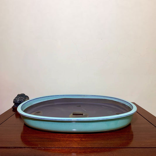 A low, oval glazed pot with a light blue rim and dark grey interior sits on a wooden surface.