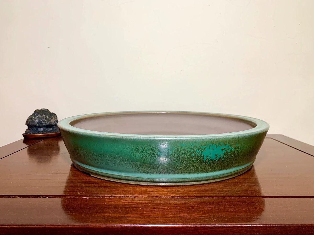 A low, wide ceramic pot with a glossy green glaze and speckled texture sits on a wooden surface.