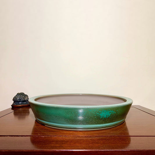 A low, wide ceramic pot with a glossy green glaze and speckled texture sits on a wooden surface.