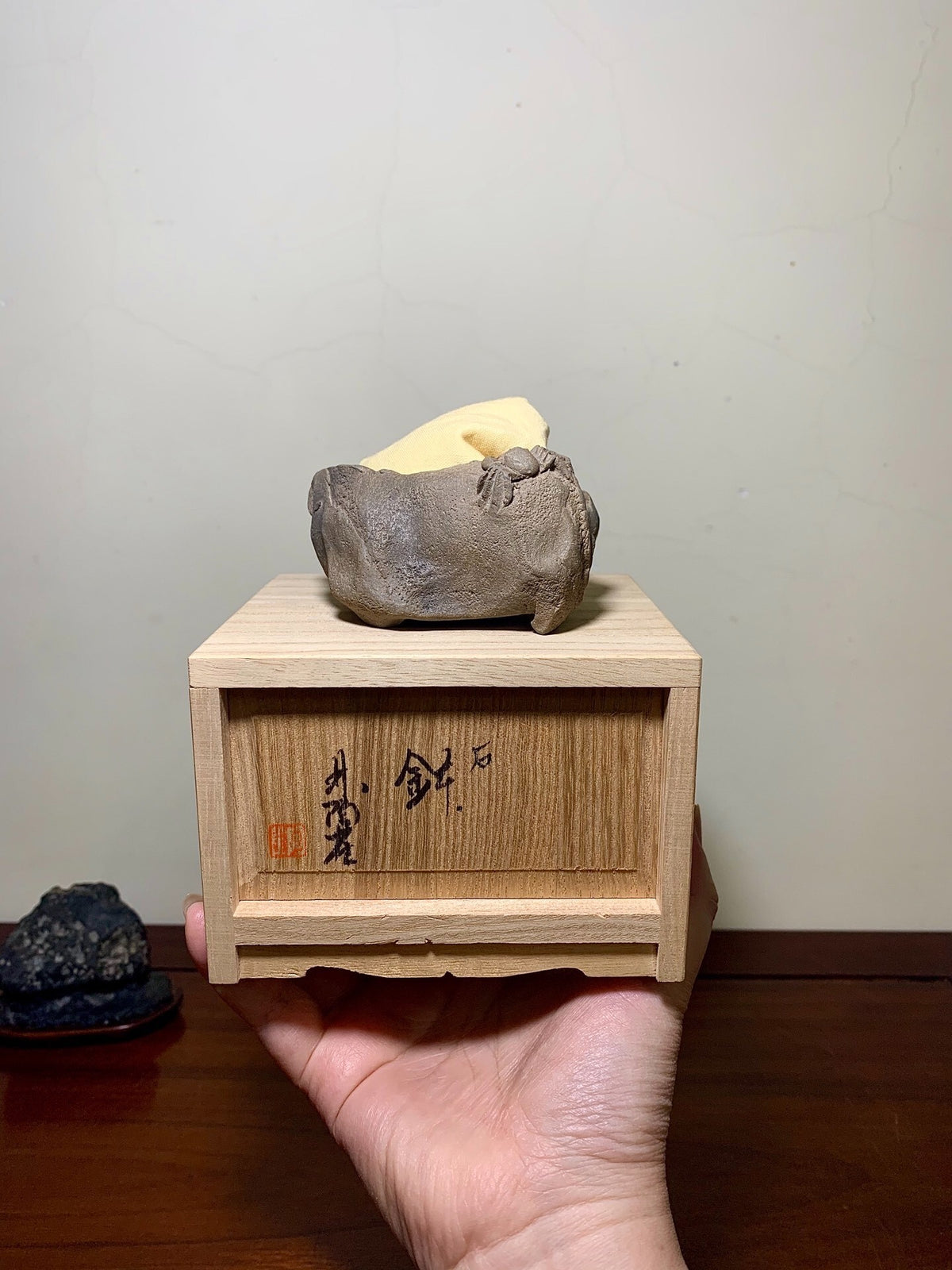 A hand holds a wooden box with Japanese characters, topped by a grey, unglazed clay pot.