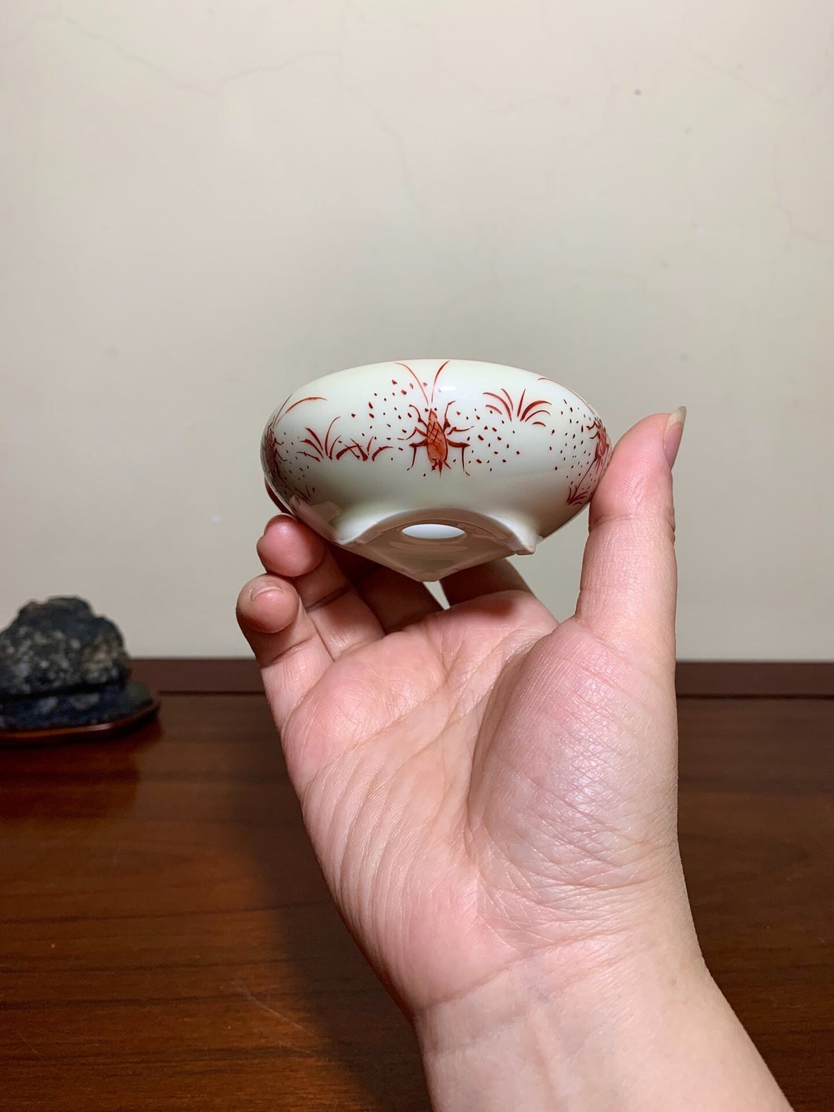 A hand holds a small, white, glazed pot with red floral patterns and a central hole.