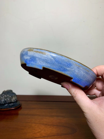 A hand holds a glazed ceramic pot with a blue and gold speckled glaze and a brown rim.