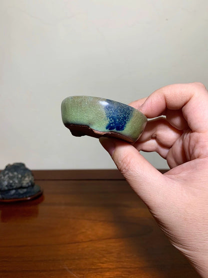 A hand holds a small, glazed ceramic bowl with green and blue glaze, unglazed rim.