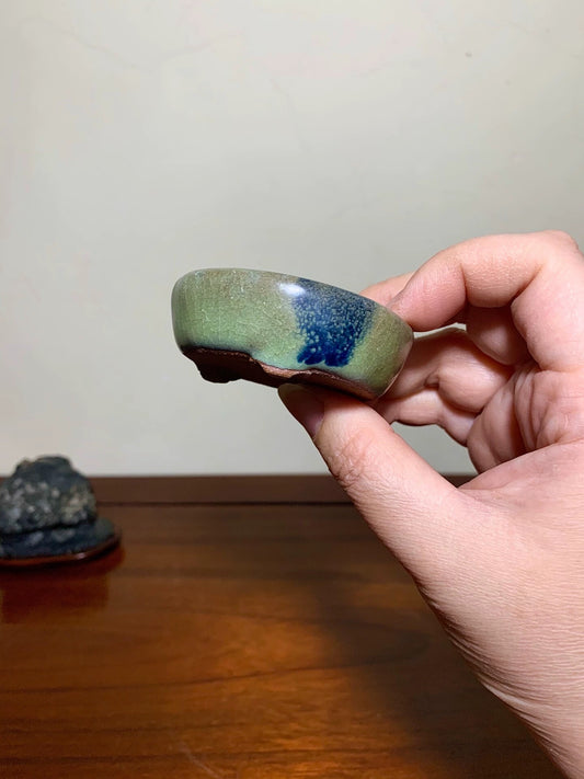 A hand holds a small, glazed ceramic bowl with green and blue glaze, unglazed rim.