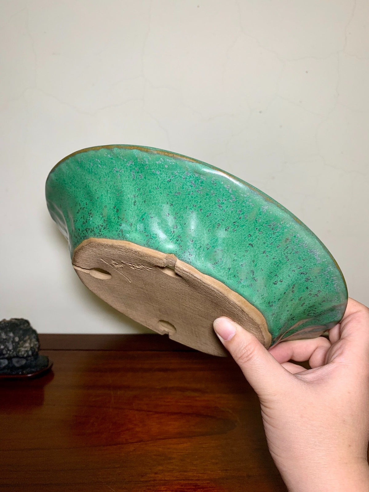 A hand holds a glossy green glazed pot with a brown unglazed base and two drainage holes.