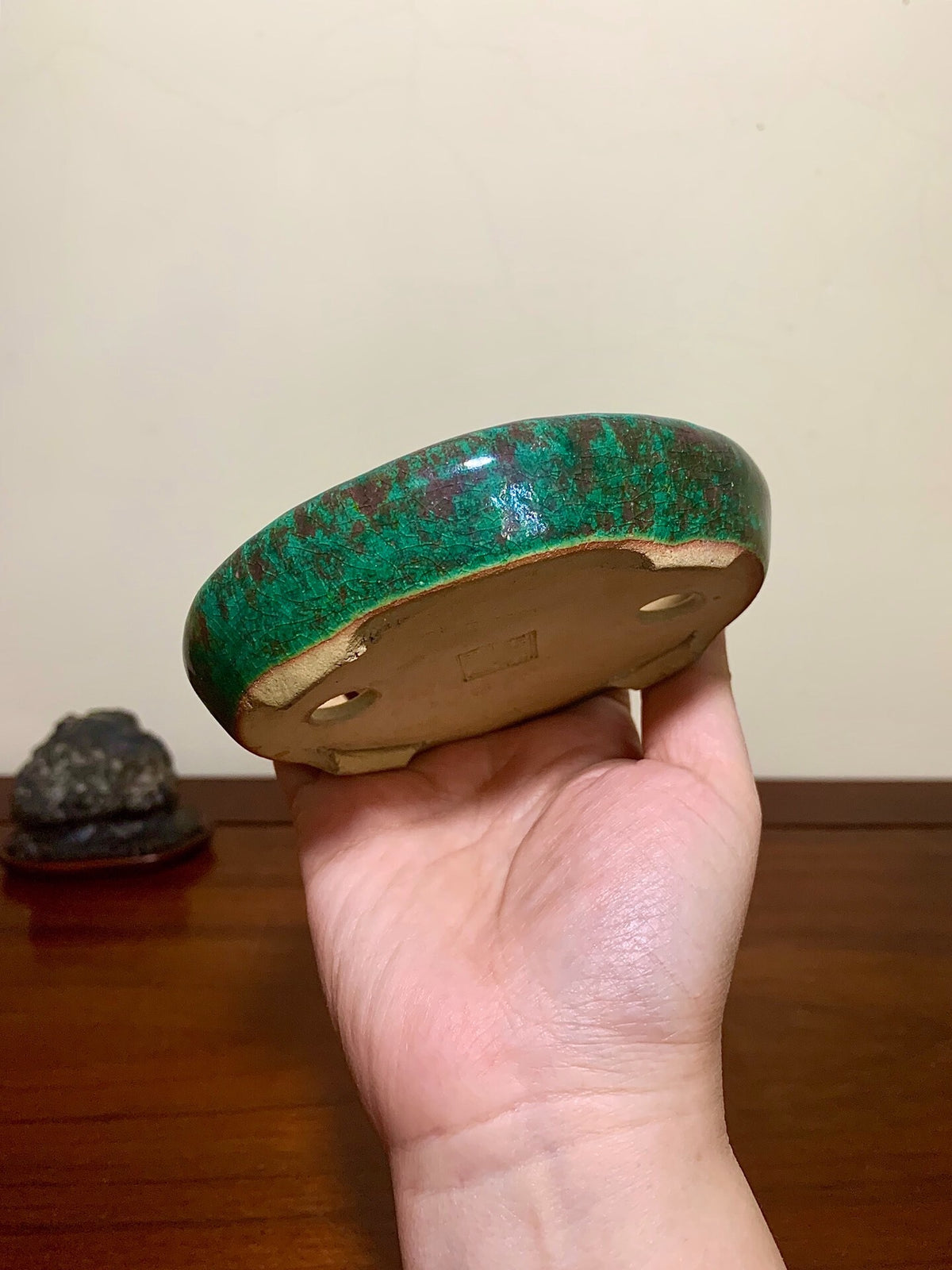 A hand holds a small, round, glossy green glazed pot with brown speckles and unglazed rim.
