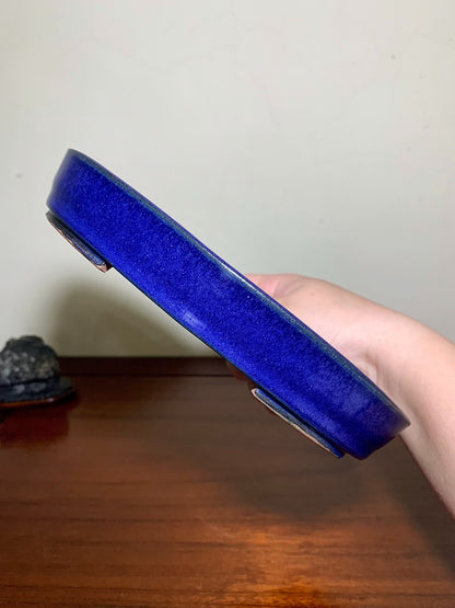 A hand holds a glossy, deep blue ceramic pot with a wide rim and two small metal feet.