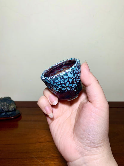 A hand holds a small, glossy ceramic pot with a blue and black speckled glaze.