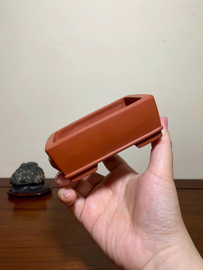 A hand holds a terracotta rectangular bonsai pot with a glossy finish and unglazed rim.