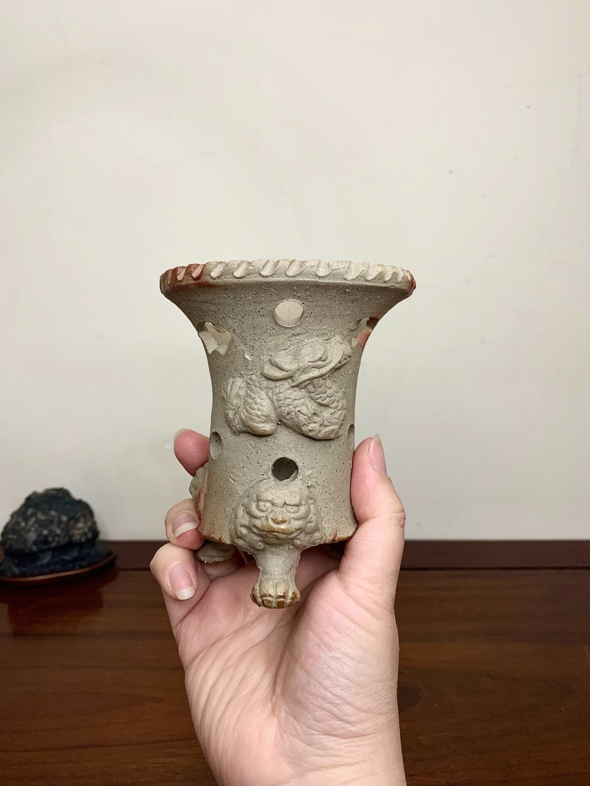 A hand holds a beige, glazed ceramic pot with dragon relief and three feet, rimmed with ridges.