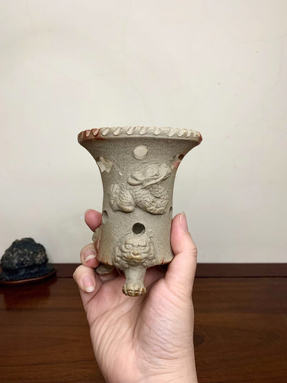 A hand holds a beige, glazed ceramic pot with dragon relief and three feet, rimmed with ridges.