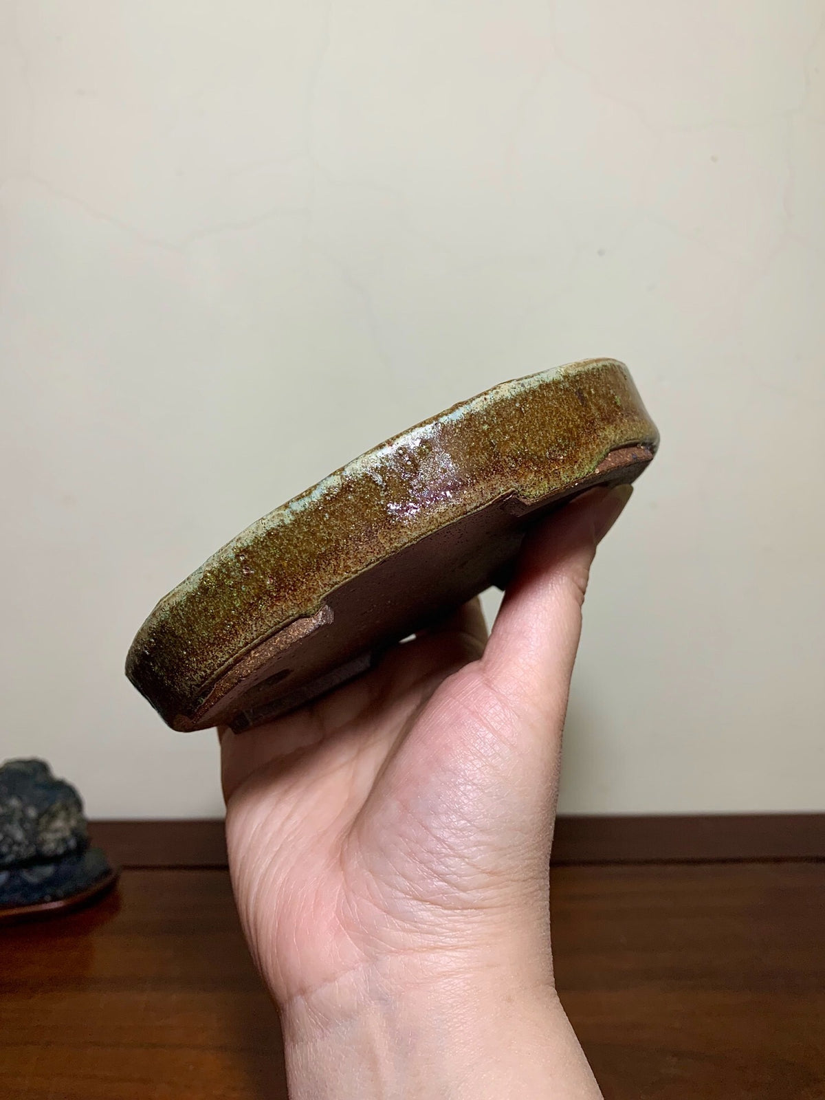 A hand holds a small, round, glazed ceramic pot with a mottled brown and green glaze.