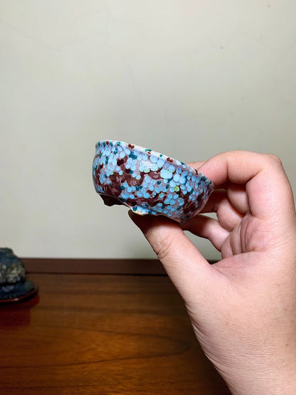 A hand holds a small, shallow bowl with a glossy, speckled blue and brown glaze.