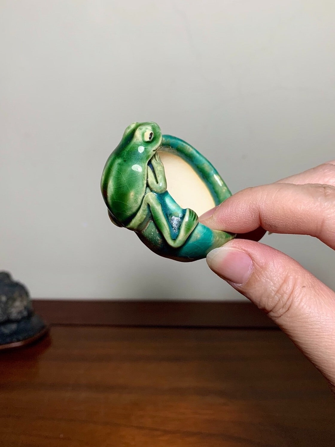 A hand holds a small, glossy green ceramic frog-shaped pot with a glazed interior.