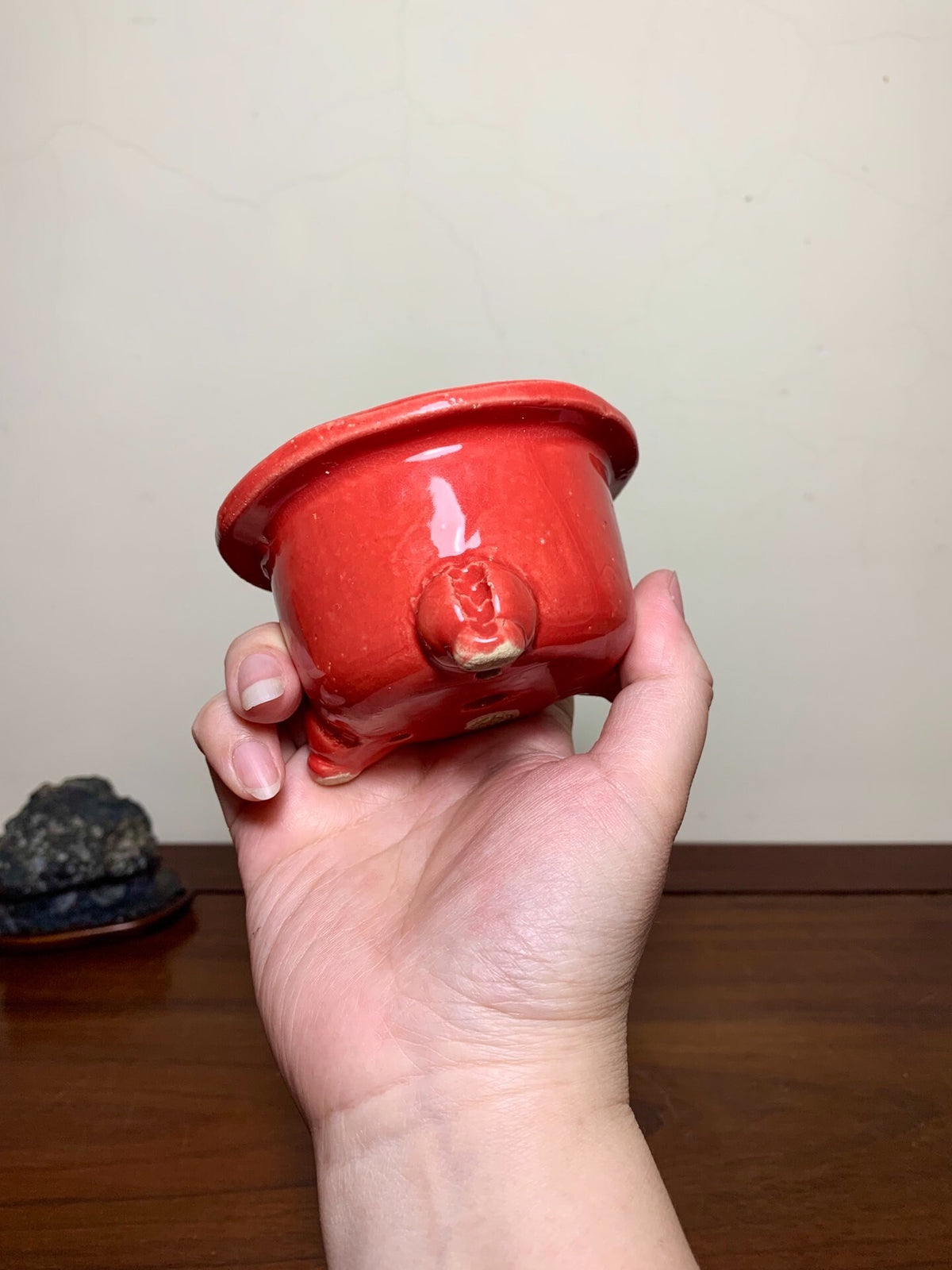 A hand holds a glossy, bright red ceramic pot with three feet and a small spout.