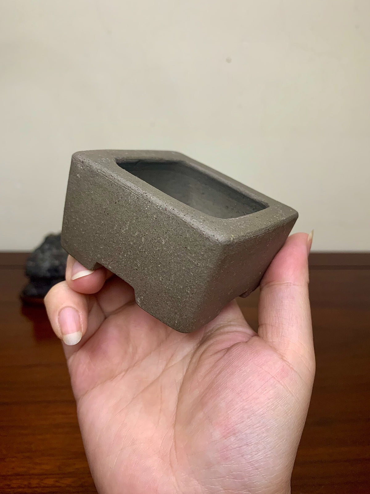 A hand holds a small, grey, unglazed square pot with a matte finish and a rectangular opening.