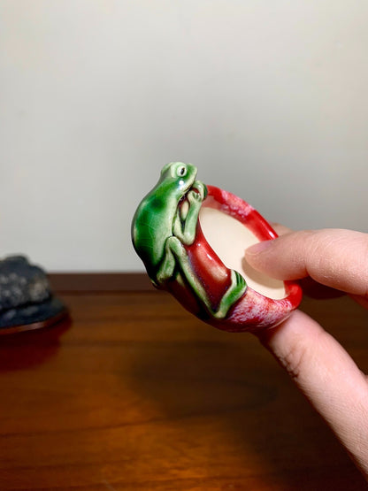 A hand holds a small, glossy red pot with a green frog decoration on its rim.