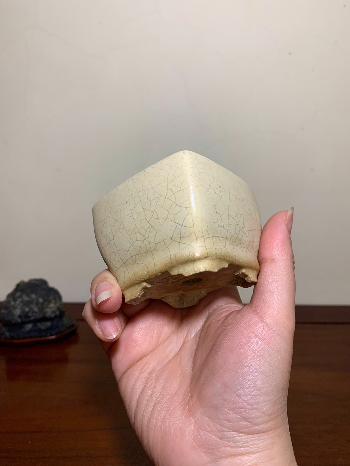 A hand holds a small, cream-glazed, hexagonal pot with a crackled surface and unglazed base.