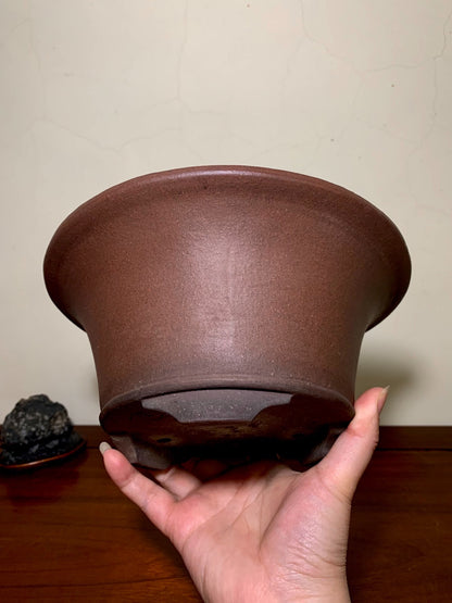 A hand holds a matte brown unglazed ceramic pot with a wide rim and flared base.