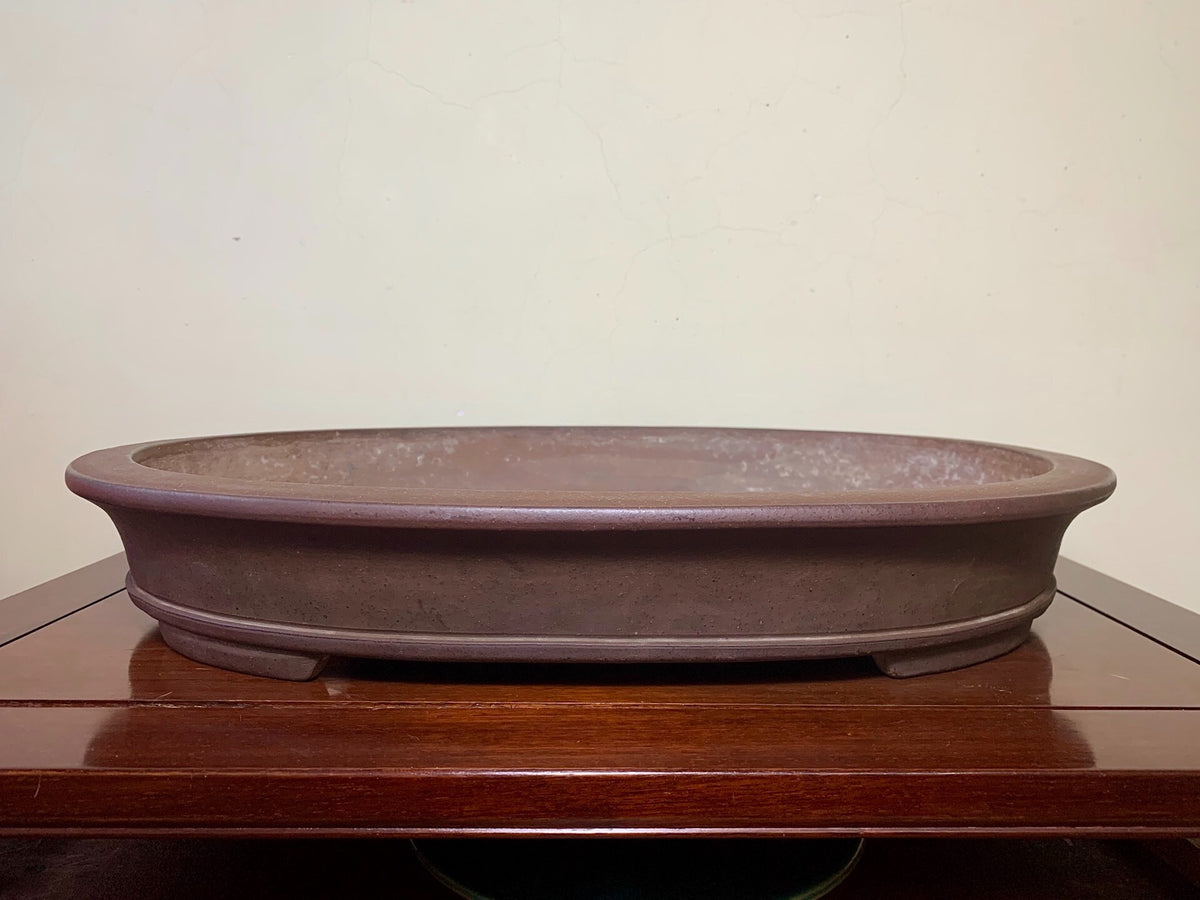 A low, oval, unglazed brown ceramic pot with a matte finish and subtle surface texture.