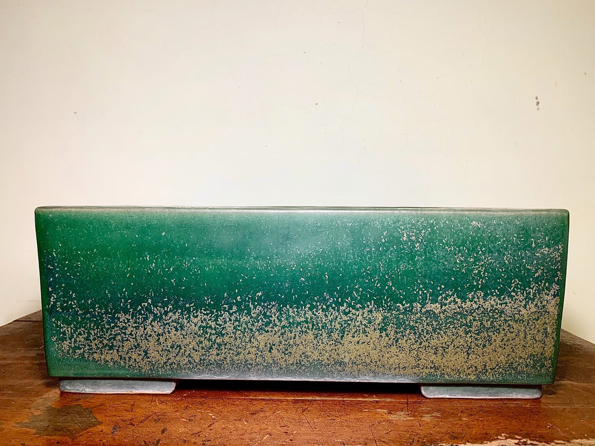 A long, rectangular green glazed pot with a speckled lower half rests on two grey feet.