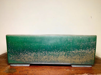 A long, rectangular green glazed pot with a speckled lower half rests on two grey feet.