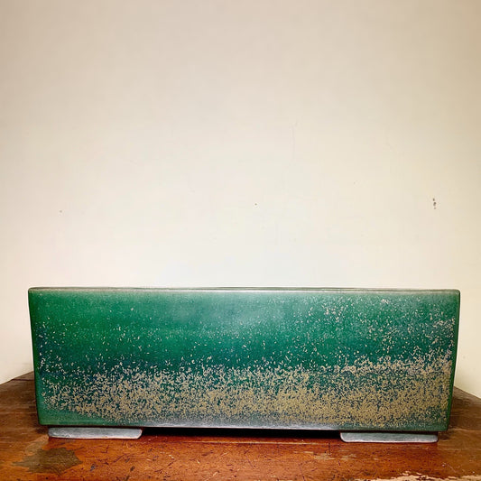 A long, rectangular green glazed pot with a speckled lower half rests on two grey feet.