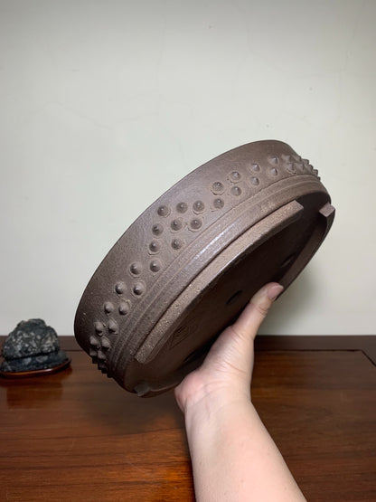 A hand holds a brown, unglazed ceramic pot with raised dots and ridges around its rim.