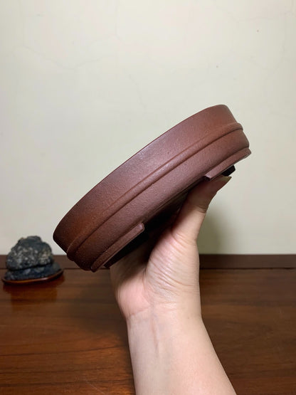 A hand holds a brown, unglazed ceramic pot with a wide rim and subtle ridges.