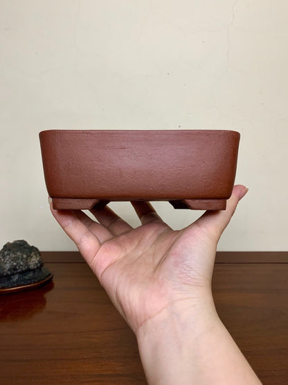 A hand holds a small, unglazed reddish-brown rectangular pot with a matte finish and four short feet.