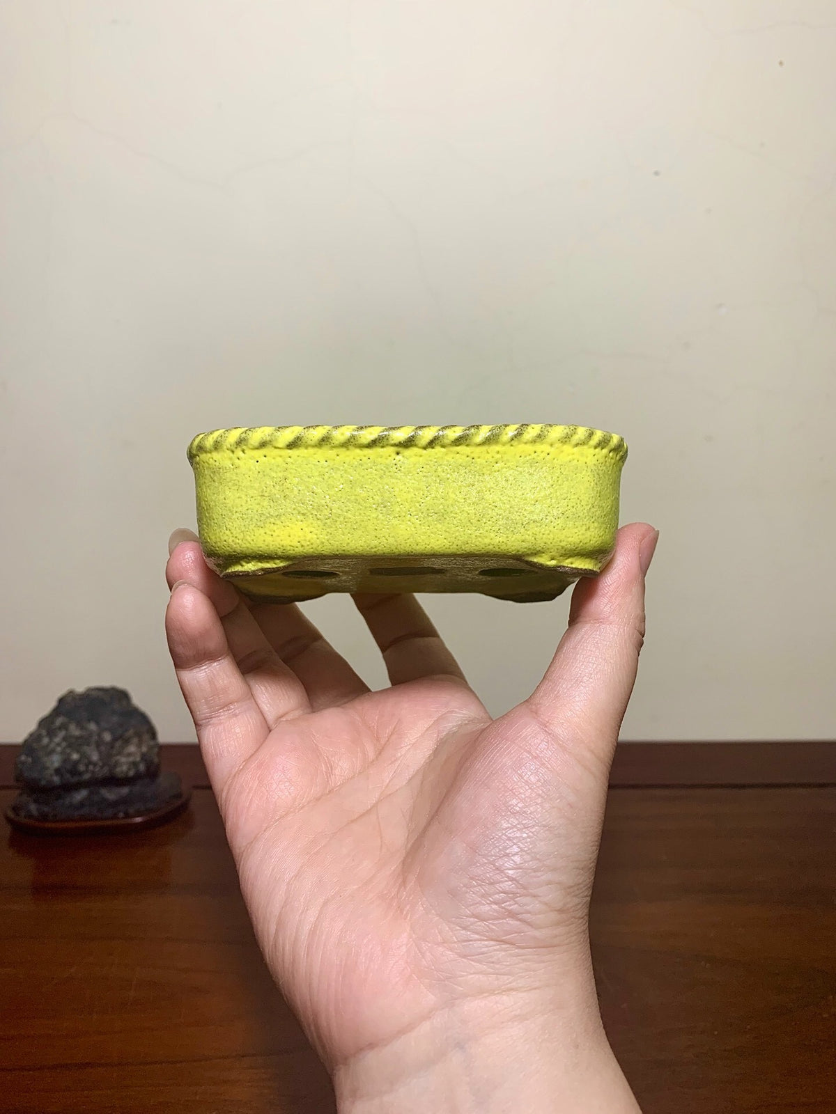 A hand holds a bright yellow, rectangular pot with a textured, rope-like rim and four drainage holes.