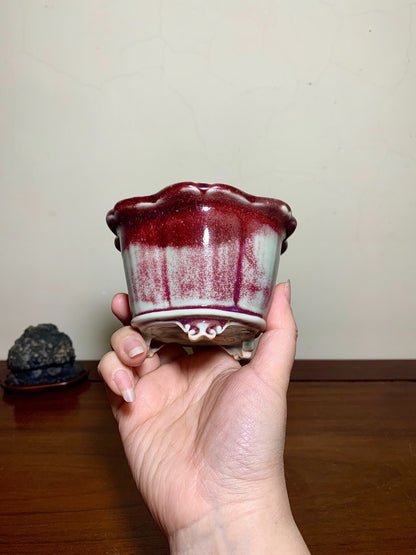 A hand holds a small, glossy ceramic pot with a flared rim, glazed in deep red and pale green.