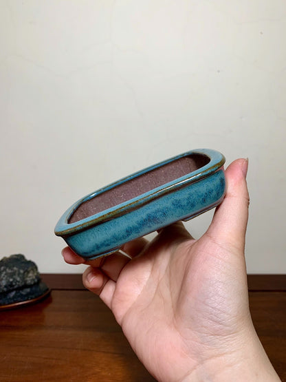 A hand holds a small, rectangular, blue-glazed ceramic pot with a brown interior.