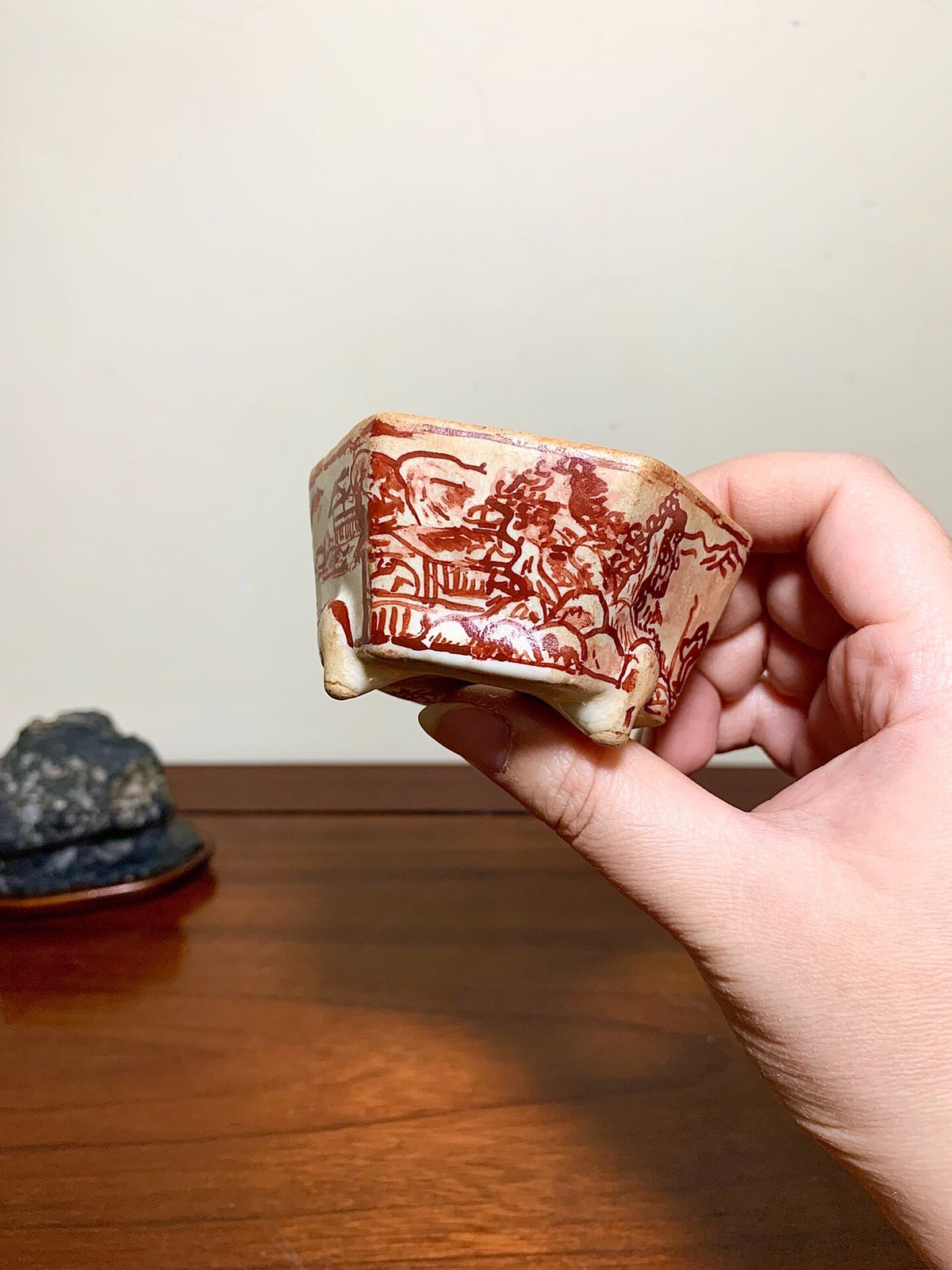 A hand holds a small, hexagonal, cream-glazed pot with reddish-brown landscape decoration.