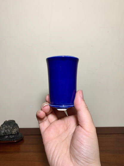 A hand holds a small, glossy cobalt blue ceramic cup with a flared rim and unglazed base.