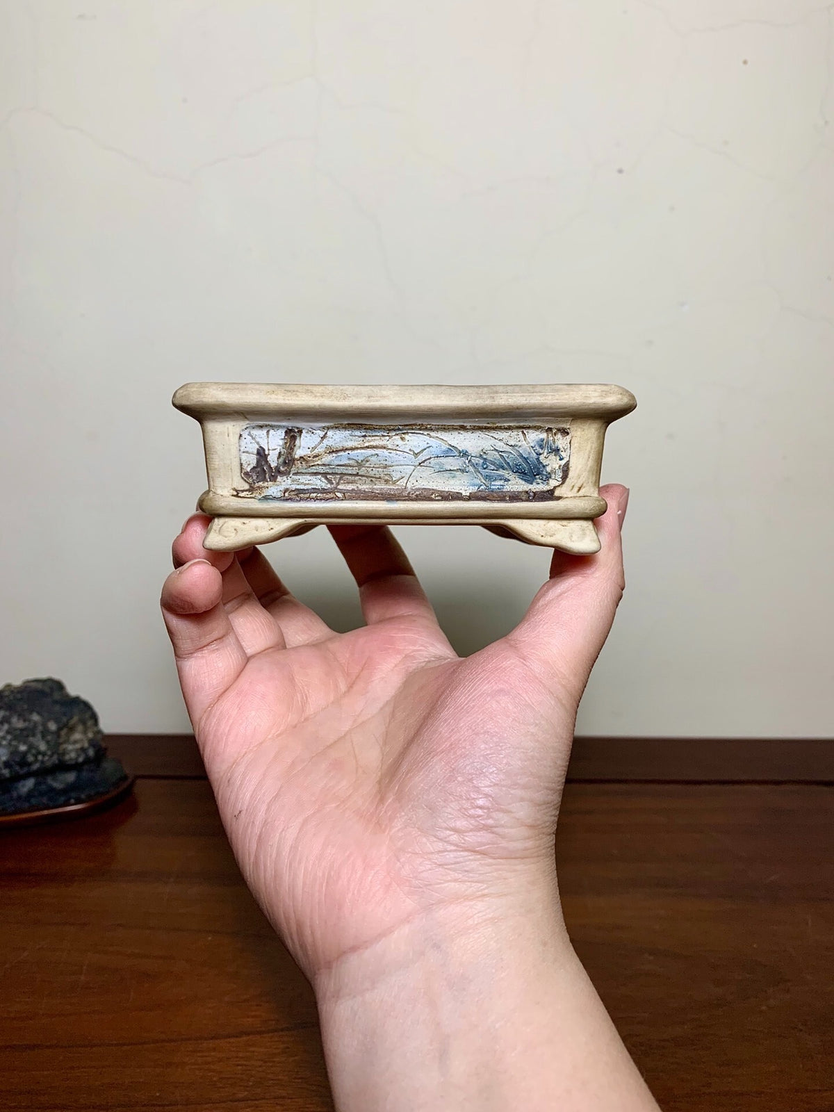 A hand holds a small, rectangular, beige-glazed pot with a blue and white painted landscape scene.