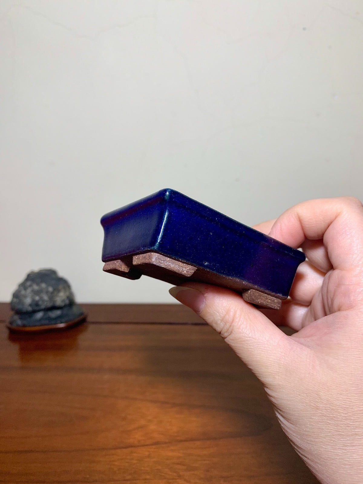 A hand holds a small, glossy cobalt blue rectangular pot with unglazed brown feet.