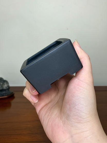 A hand holds a small, matte black square pot with a shallow, unglazed interior.