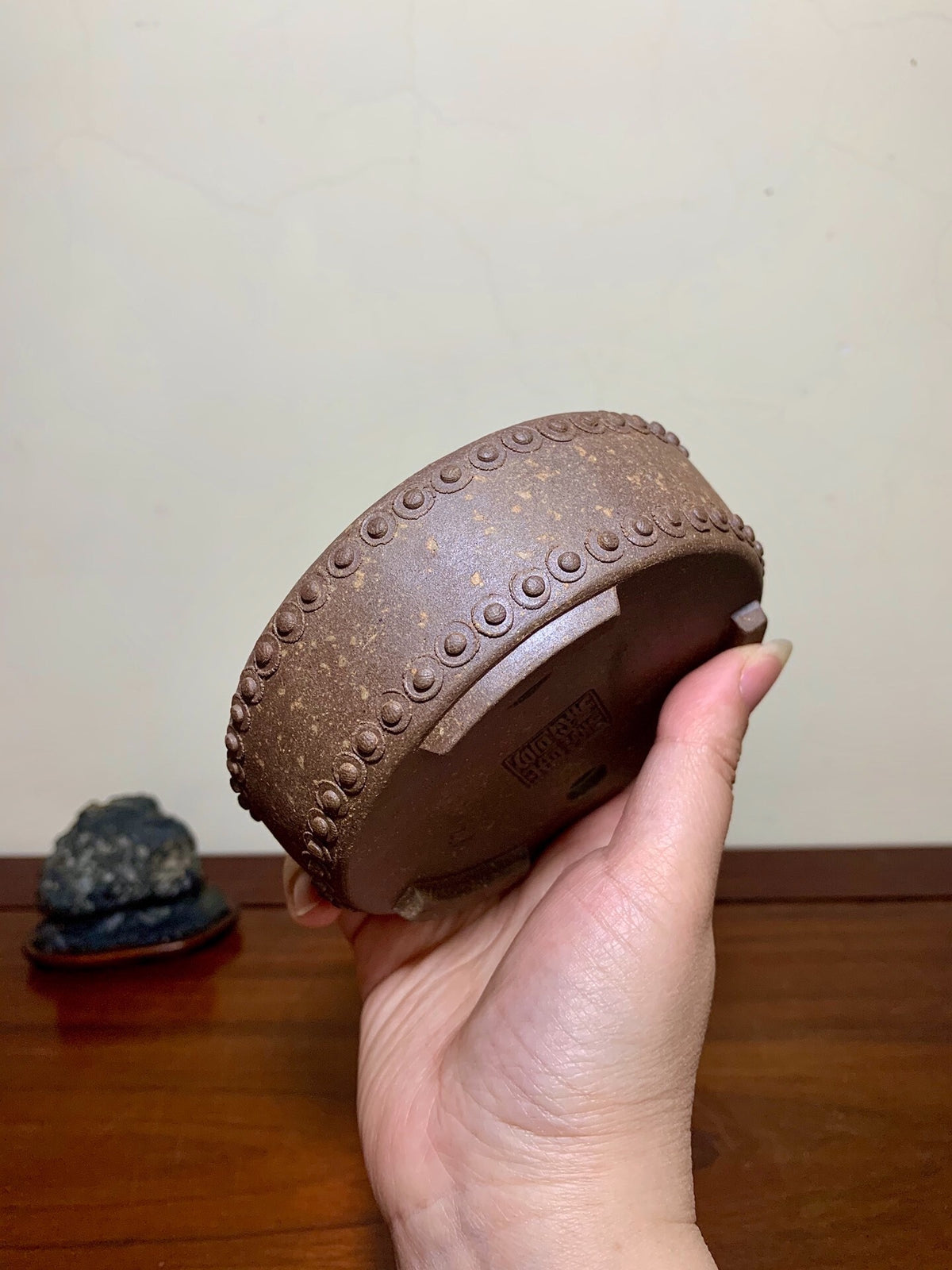 A hand holds a brown, unglazed ceramic pot with a beaded rim and visible maker's mark.