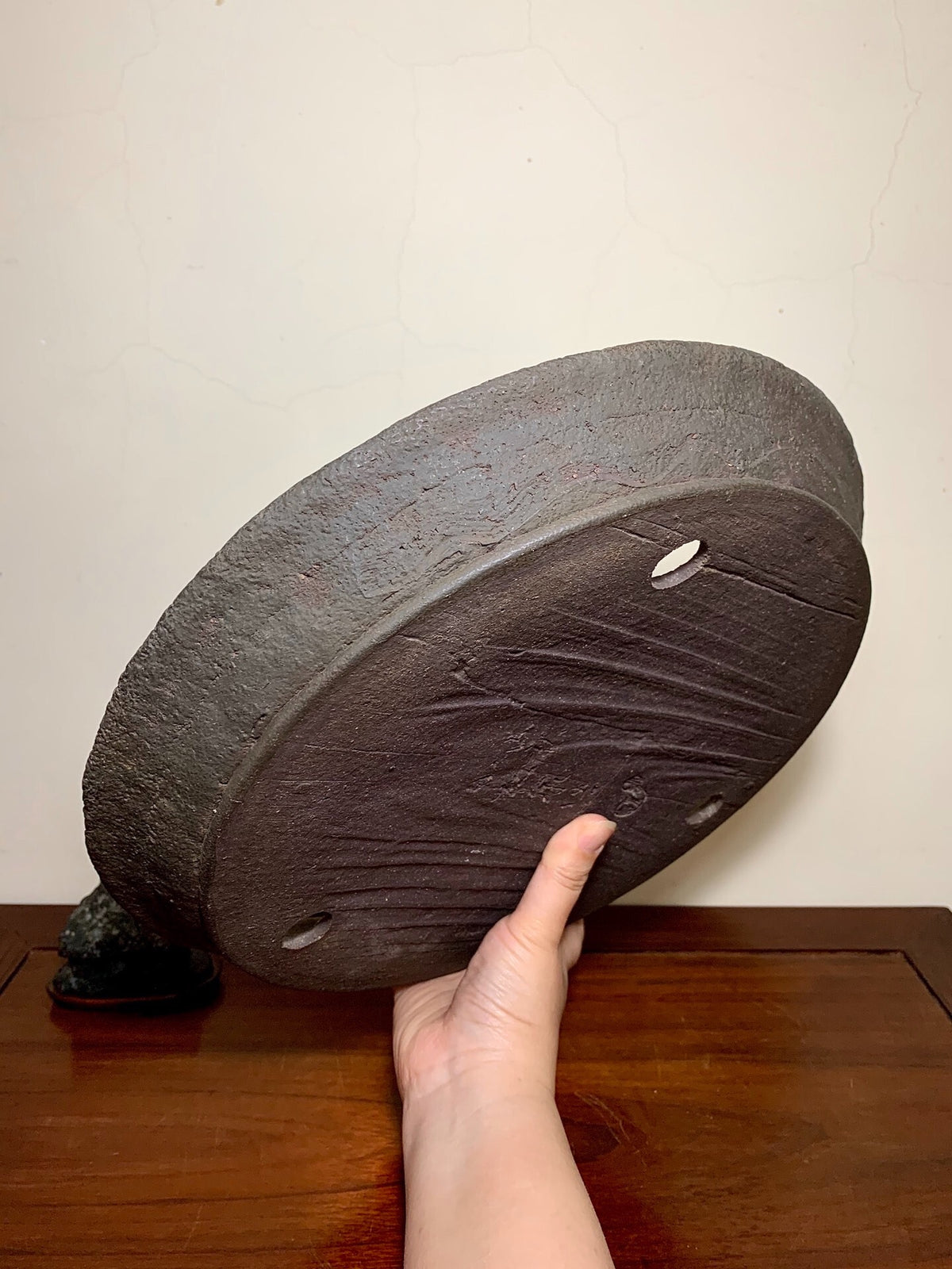 A hand holds a dark, textured, unglazed ceramic pot base with three drainage holes and incised markings.