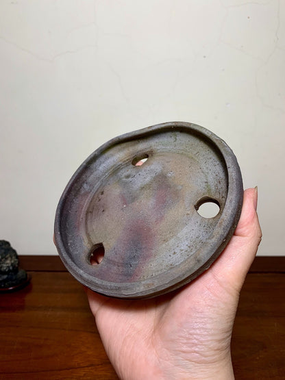 A hand holds a dark, glazed ceramic pot base with three holes and a mottled surface.