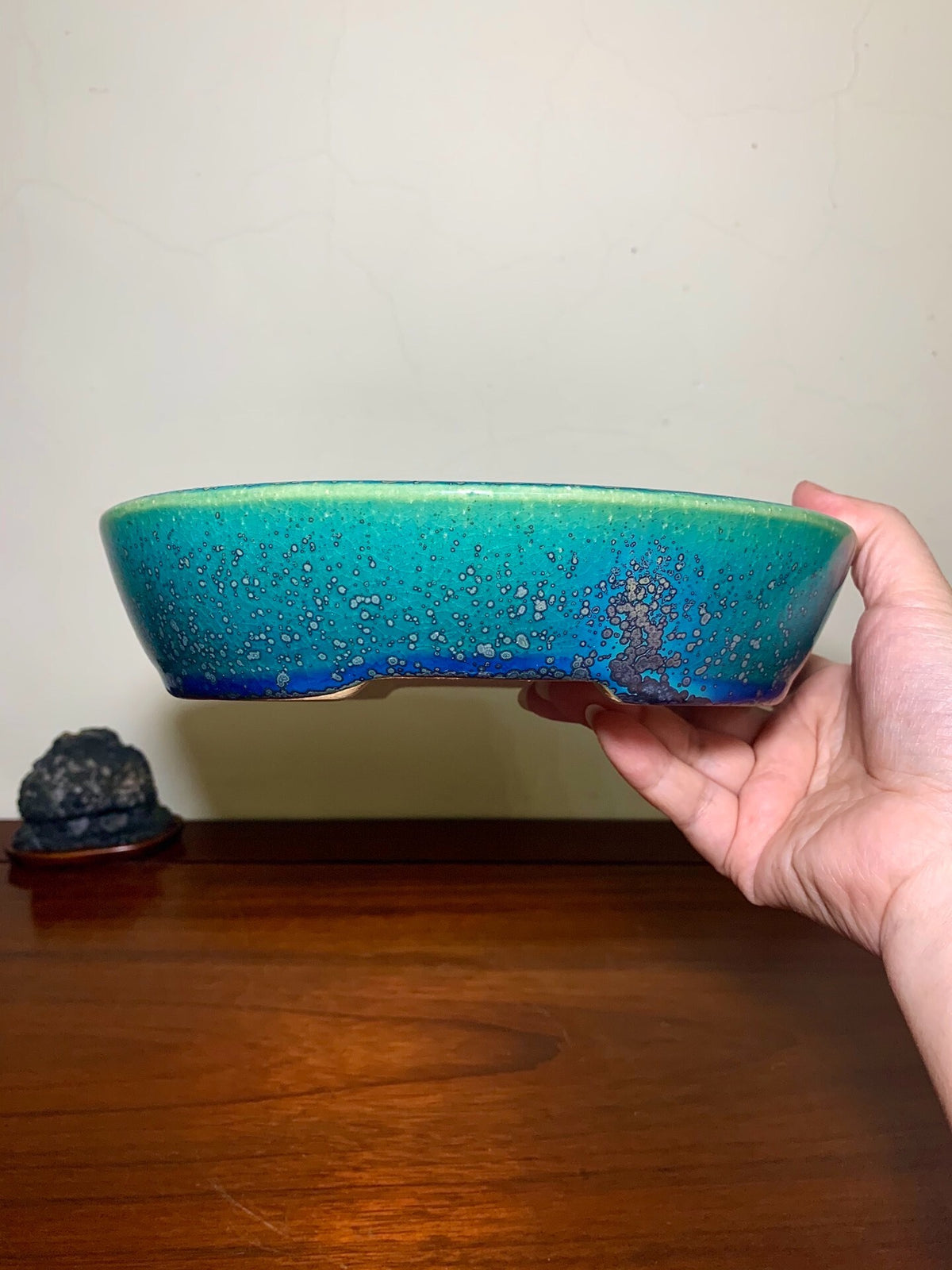 A hand holds a glossy, oval-shaped pot with a gradient turquoise and blue glaze speckled with white.