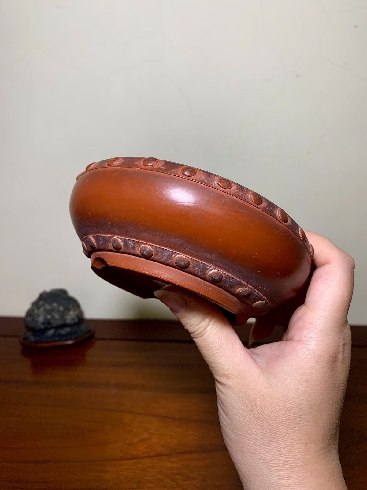 A hand holds a glossy, rounded brown pot with raised dot decorations around its rim and base.