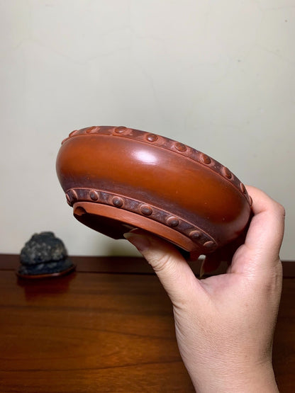A hand holds a glossy, rounded brown pot with raised dot decorations around its rim and base.