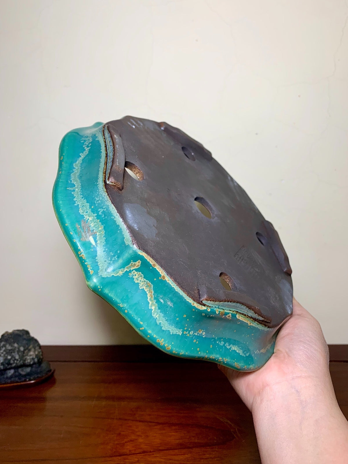 A hand holds a glazed ceramic pot with a teal rim and dark brown base, showing drainage holes.