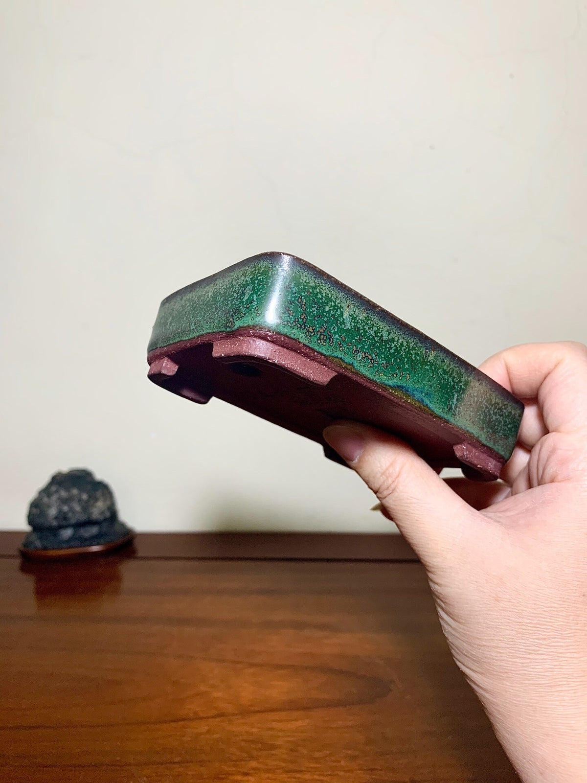 A hand holds a rectangular glazed pot with a green and brown speckled glaze.