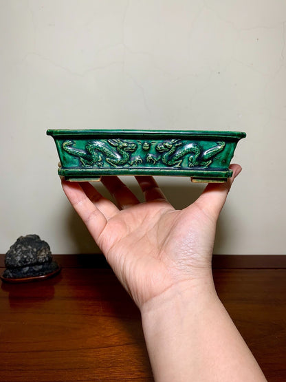 A hand holds a green glazed rectangular pot with raised dragon decoration.