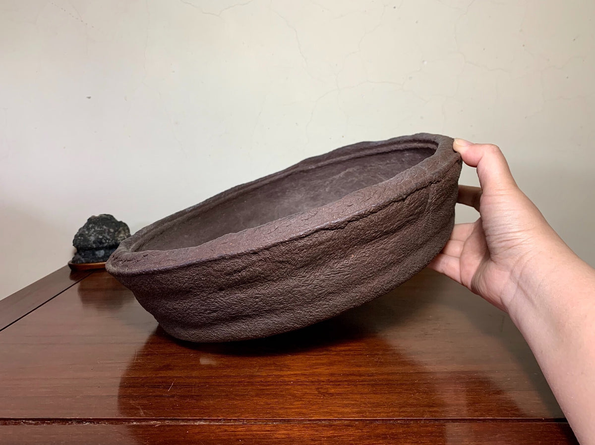 A hand holds a dark brown, textured, unglazed ceramic bowl with a wide rim and shallow depth.