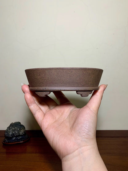 A hand holds a small, round, unglazed brown ceramic pot with three feet.