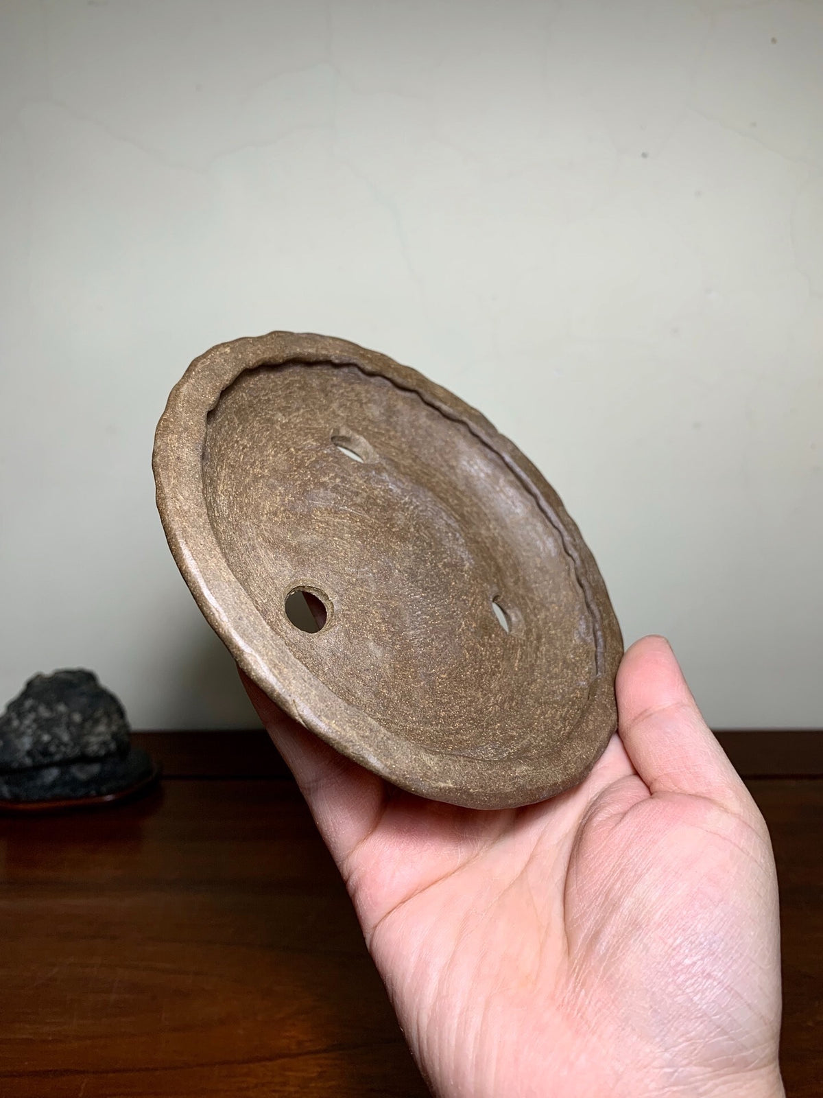 A hand holds a brown, unglazed ceramic pot lid with a central hole and two smaller holes.